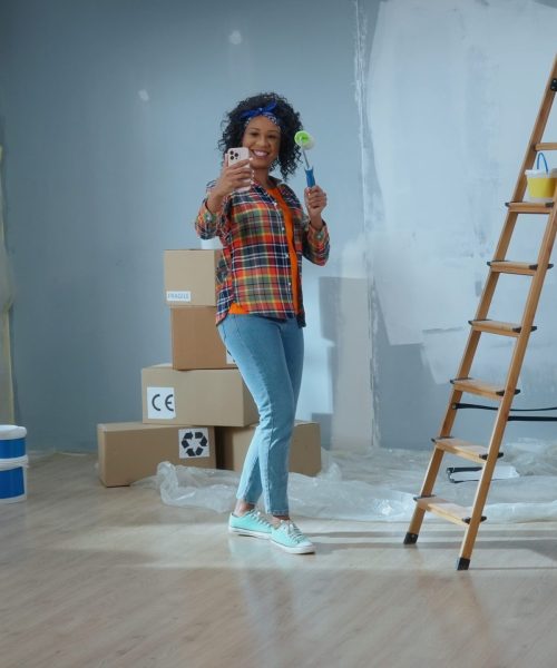 Young African American woman is making video call using her mobile phone, smiling and showing a paint roller. Black female in checkered shirt communicates remotely through camera. Concept of repair, online communication, modern technology.